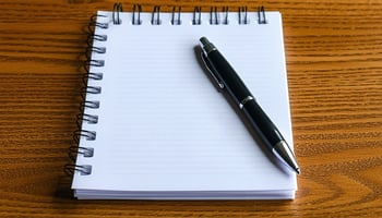 A notepad with blank pages and a pen placed on a wooden table, suggesting a workspace for writing or brainstorming.