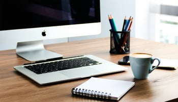 The image features a professional-looking office space with a sleek wooden desk, adorned with a laptop, notepad, and a steaming cup of coffee. 