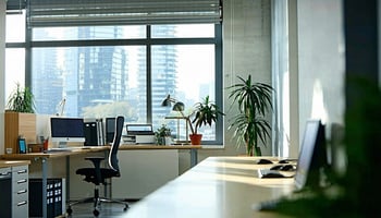 A modern office environment featuring desks with computers, office supplies, plants, and large windows offering a view of a city skyline.