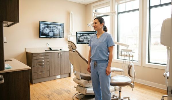 Dentist in light blue scrubs standing in a modern dental operatory beside a dental chair with panoramic X-ray images on wall monitors