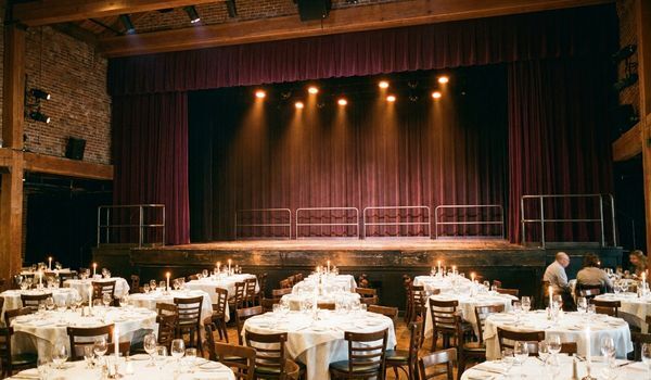 Interior of an elegant dinner theater with round linen-covered tables, candles, and a stage with burgundy velvet curtains under warm spotlights