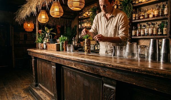 A bartender in a linen shirt garnishing a ceramic tiki mug behind a carved wooden tiki bar with bamboo pendant lights and rum bottle shelves