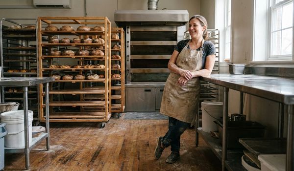 Bakery owner in a flour-dusted apron standing in a commercial bakery kitchen beside wooden racks of fresh artisan bread and a deck oven
