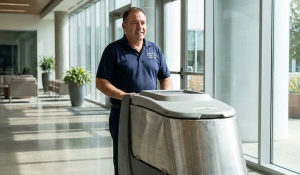 Commercial cleaning business owner in a navy uniform standing beside a walk-behind floor scrubber in a bright corporate office lobby