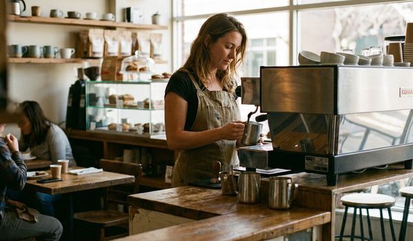 Coffee shop owner in a canvas apron steaming milk at a commercial espresso machine inside an independent cafe