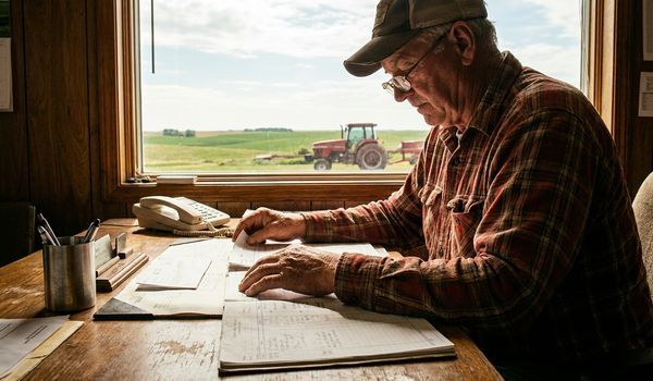 American farmer reviewing financial paperwork at a wooden desk in a farm office with green fields and a tractor visible through the window