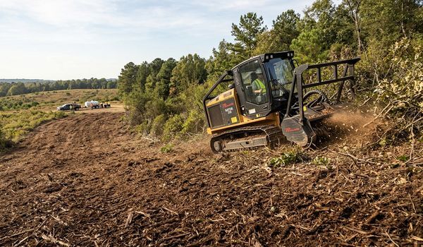 Tracked forestry mulcher clearing brush and small trees on a commercial land clearing job site, with an operator in the cab and support crew nearby