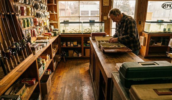 Owner of a family bait and tackle shop arranging tackle trays at a wooden counter, with fishing rods, lure displays, and live bait tanks in the background