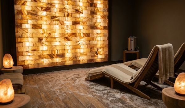Interior of a salt therapy spa with a glowing backlit Himalayan salt brick wall, lounge chairs, and carved salt lamps