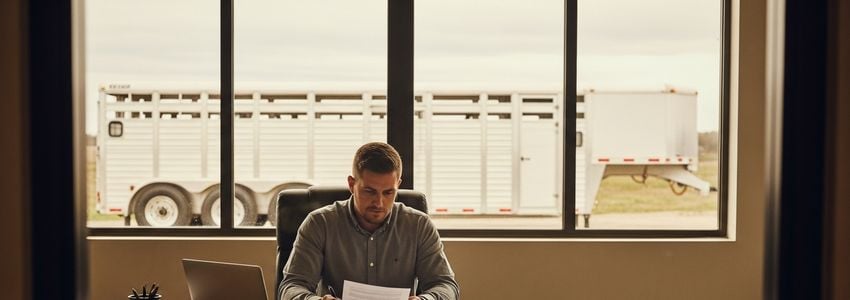 A farm business owner reviewing livestock trailer financing documents at an office desk with a livestock trailer visible through the window